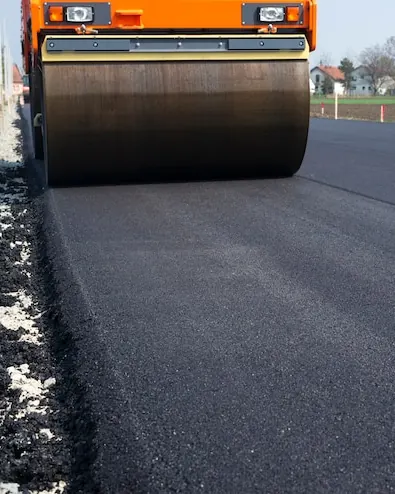 Cilindru compactor pe asfalt proaspăt, cu casele unui sat în spate